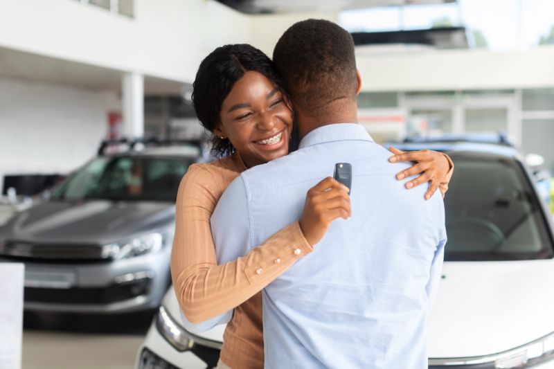 A young woman excitedly hugs her boyfriend while holding car keys. There are two cars in the background.